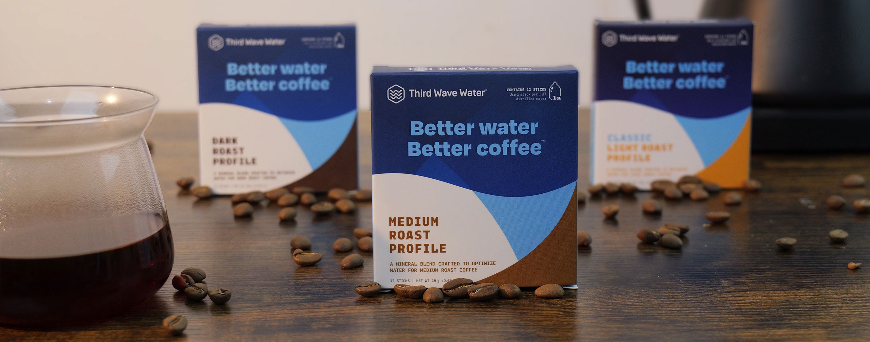 Three boxes of Third Wave Water with a glass of coffee and coffee beans on a wooden surface.