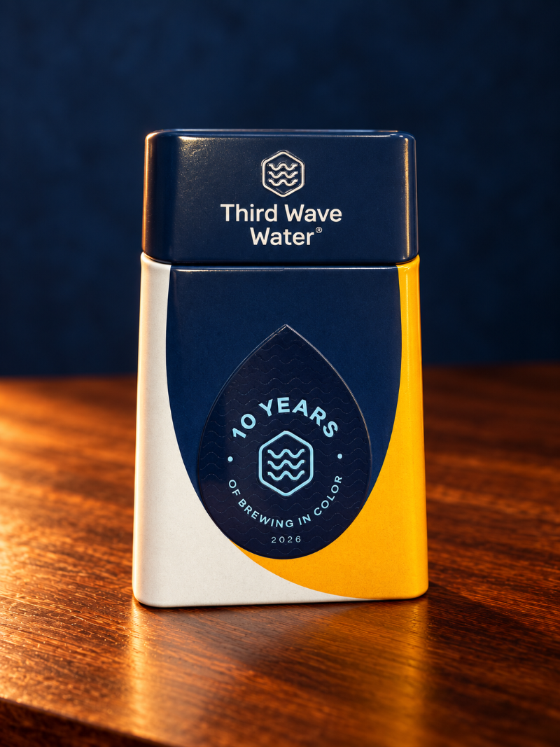 A rectangular metal tin of Third Wave Water coffee on a wooden table against a dark blue background. The tin has a dark blue top and front label, a white side panel, and a yellow lower-right section.